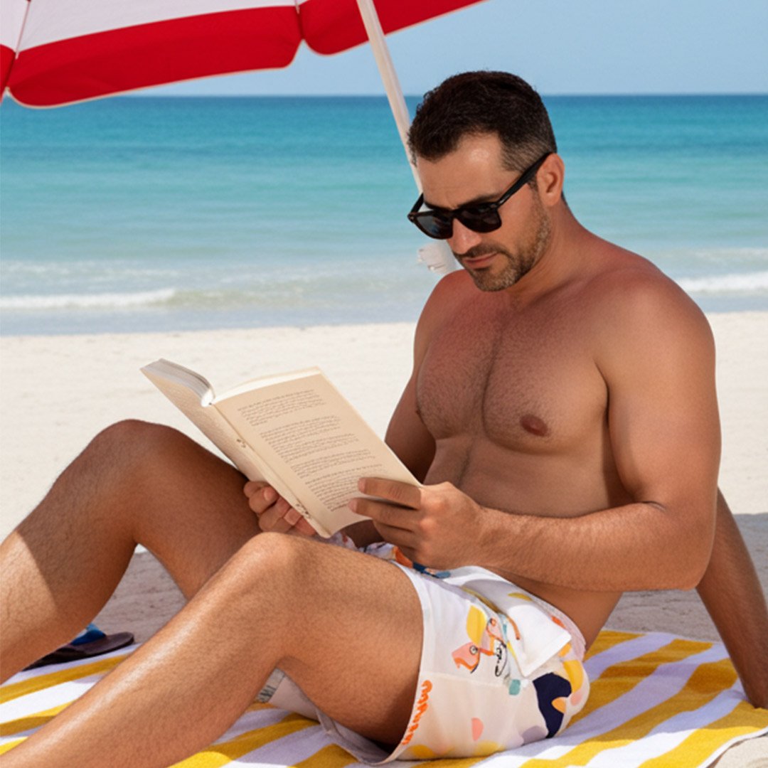 Man sitting on the beach under an umbrella, reading a book while wearing colorful Bénieller swim trunks.